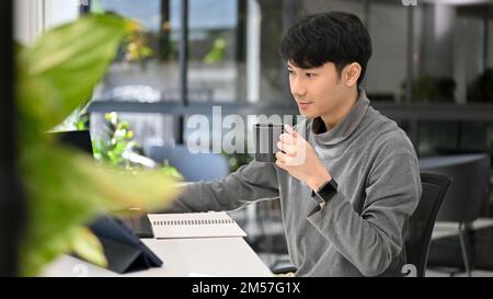 Focused young Asian male graphic designer or website developer sipping coffee while working at his office desk in the office. Stock Photo