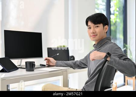 Handsome and smart young Asian male graphic designer or program developer sits at his desk in modern office, smiling and looking at the camera. Stock Photo