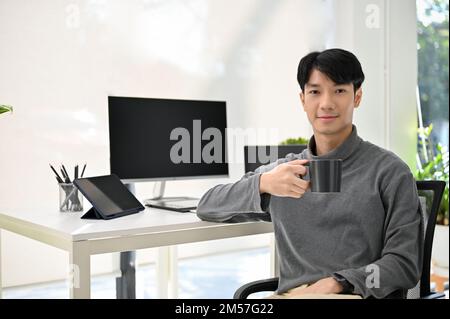 Handsome and successful Asian male developer or manager sits at his desk, holding a cup of coffee, smiling and looking at the camera. Stock Photo