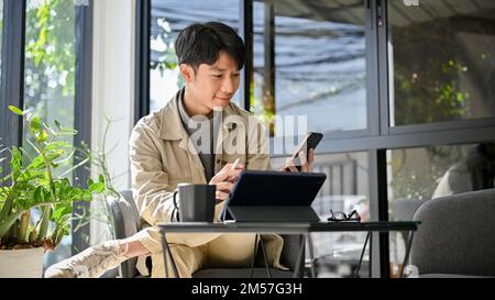 Happy and relaxed young Asian an using his phone and portable tablet at the coffee shop. remote working concept Stock Photo