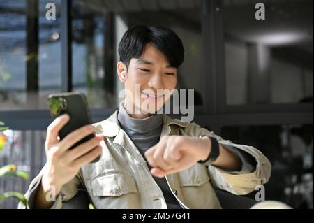 Smiling and happy millennial Asian man in casual clothes holding his phone, checking time on his smartwatch while relaxing in the coffee shop. Stock Photo
