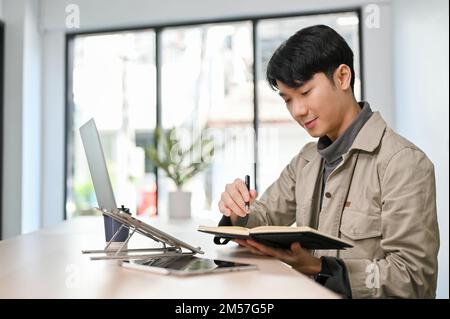 Good looking young Asian male office worker remote working at the coffee shop co-working space, checking his meeting schedule on his planner book. Stock Photo