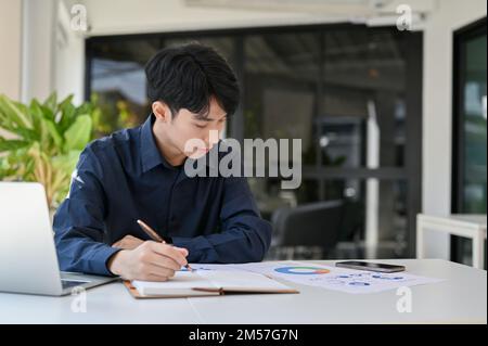 Focused young Asian male financial analyst or accountant working on financial report, analyzing financial data, reviewing sales report. Stock Photo