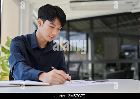 Concentrated young Asian male financial analyst or accountant working on financial report, analyzing financial data on the report. Stock Photo