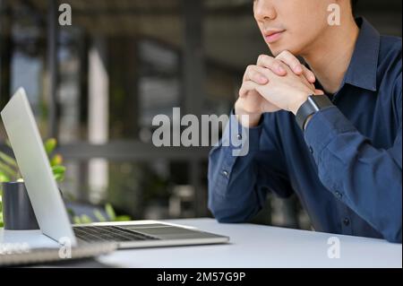 Focused and thoughtful young Asian businessman or male office worker pensively thinking about his project, sits in front of his laptop computer. Stock Photo