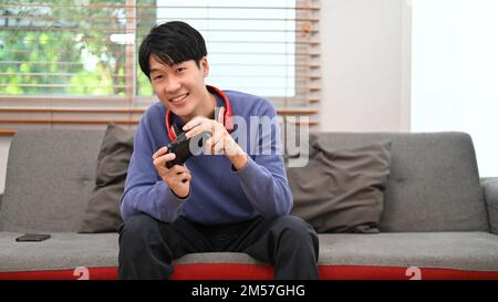 Joyful and happy young Asian man in casual clothes enjoys playing video game alone in his living room. Stock Photo