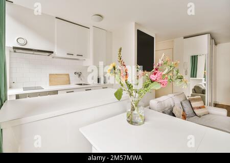 some flowers in a vase on a kitchen table with white cupboards and green drapes behind the counter area Stock Photo