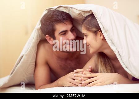 Speaking with their eyes. a loving young couple smiling at each other while lying under a blanket together. Stock Photo