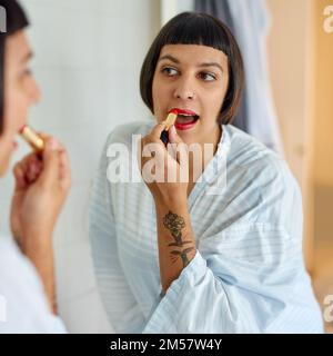 This will make my lips pop. a young woman in a bathrobe applying red lipstick in her bathroom mirror in the morning. Stock Photo