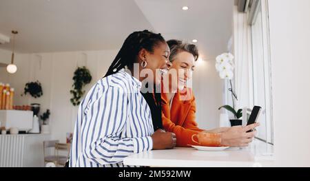 Smiling business women video calling their colleagues on a smartphone in a coffee shop. Two happy business women having an online morning meeting with Stock Photo