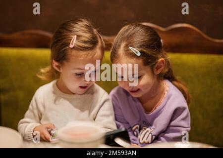 Sisters kids hold mobile phone together at cozy cafe and having fun. Stock Photo