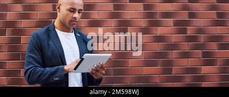 Using gadget. Young guy standing on wall browsing internet on digital tablet concentrated Stock Photo