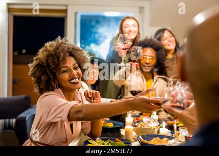 Mixed age range group of friends toasting at dinner party, focus on woman face, Brazilian people drinking red wine and eating vegan food, happy family Stock Photo