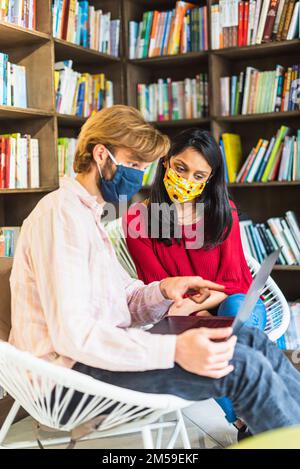 Two young students in a library with protective mask on their face to prevent Coronavirus infection, people in smart working, the new normal concept Stock Photo