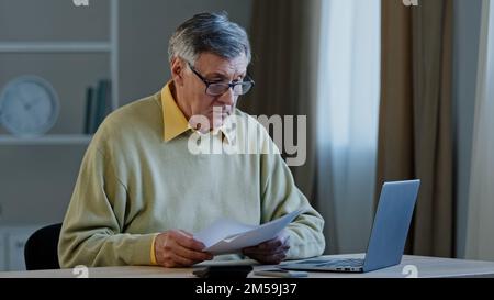 Serious old Caucasian grey-haired businessman 70s male senior mature elderly entrepreneur man sitting at table with paperwork problems with documents Stock Photo