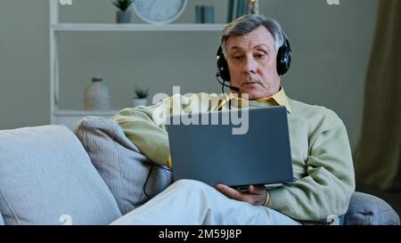 Senior 60s male on couch at home wear digital earphones talking online to laptop web camera old man using computer app for conference discuss business Stock Photo