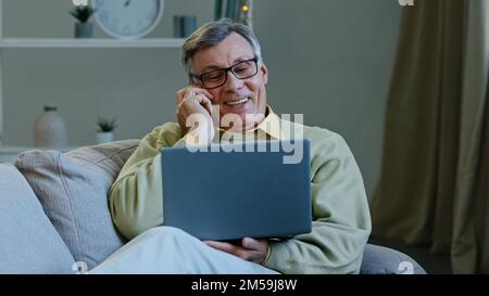 Old caucasian grandfather aged 60s man senior male with laptop sits on sofa at home talking on mobile phone makes order online book food delivery Stock Photo