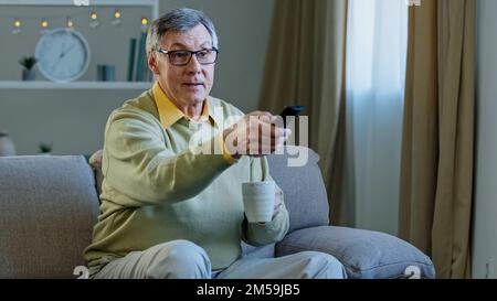 Grey-haired aged man in glasses sitting on couch watching TV enjoying relaxing at home drinking tea coffee aged male uses remote control to switch Stock Photo
