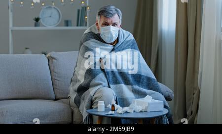 Ill sick old male in medical mask sitting on couch at home Caucasian elderly man wrapped in blanket feel unwell illness mature senior looking at Stock Photo