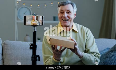 Elderly vlogger record videoblog online stream on social networks old man conducts live broadcast grandfather unpacking mystery box making video on Stock Photo