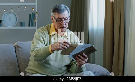 60s elderly man recipient with glasses sorting letters in living room concentrated mature grandfather sitting on sofa looking at correspondence notice Stock Photo