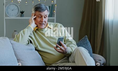 Happy elderly adult man sitting on sofa hold gadget studying app on smartphone makes online order smiling grandfather resting in room reading news Stock Photo