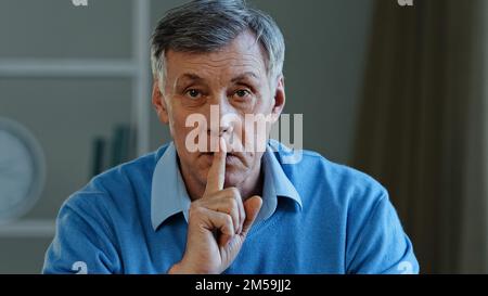 Closeup old caucasian 60s man holding index finger near mouth stop talking hush gesture silence sign elderly mature adult male telling secret looking Stock Photo