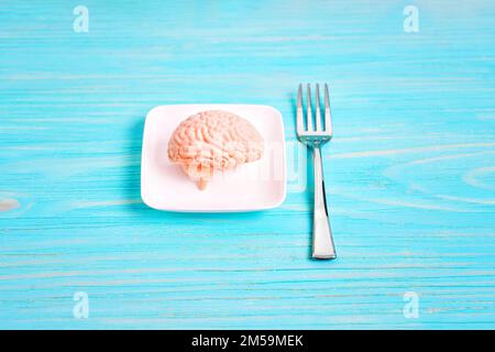 Brain-healthy dieting related concept: miniature human brain model on a tiny white plate and small silver fork isolated on blue background. Stock Photo