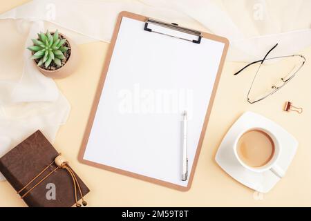 Desktop with blank clipboard, notepad, coffee cup, succulent plant and accessories on light beige background. Top view, flat lay, mockup. Stock Photo