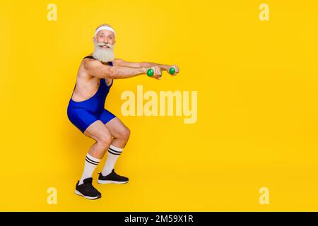 Photo of funky positive retired guy wear blue overall pumping arms sit ups empty space isolated yellow color background Stock Photo