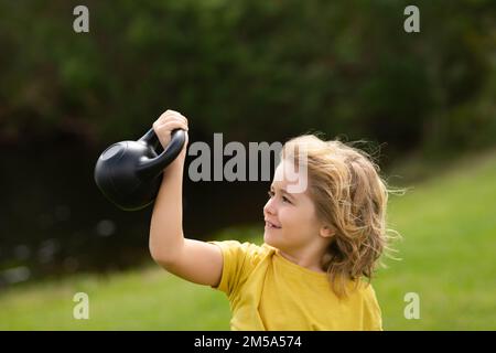 Kid lifting the kettlebell in park outside. Child boy pumping up biceps muscles with kettlebell. Fitness kids with dumbbells Stock Photo