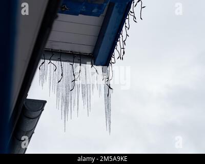 Icicles hanging on a roof gutter. Decoration lights are on the wooden roof. The sky is covered with clouds. Melt water is dripping down on a building. Stock Photo