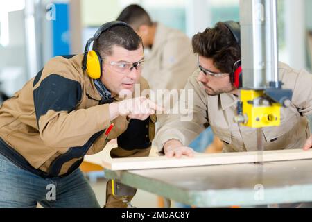experienced carpenter guiding apprentice using bench saw Stock Photo