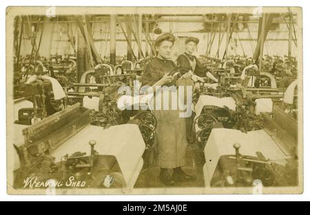 Original Edwardian era postcard of young female workers in a busy large weaving shed in front of their looms. The girl at the front appears to be of teenage years and is wearing clogs and holding a weaver's shuttle. Published by The Schofield Photographic Series circa 1906, Bollington, Cheshire, England, U.K. traditionally silk-weaving towns. Bollington is a traditionally silk weaving town in Cheshire Stock Photo