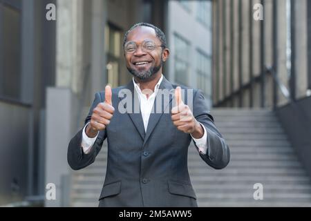 Portrait of successful African American mature businessman, senior man in business suit smiling and looking at camera showing happy thumbs up outside office building. Stock Photo