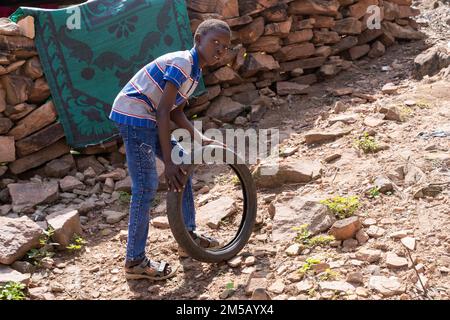 Handsome African youngster playing with his old bicycle wheel on the steep rough road in front of his house in a rural village Stock Photo
