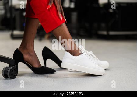 African woman changing from high heels to sneakers in the office. Tired legs.  Stock Photo
