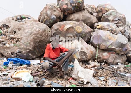 Young African boy in a red t-shirt kneeling thoughtfully in front of a huge pile of recycled plastic bottles wrapped-up in old mosquito nets in a muni Stock Photo