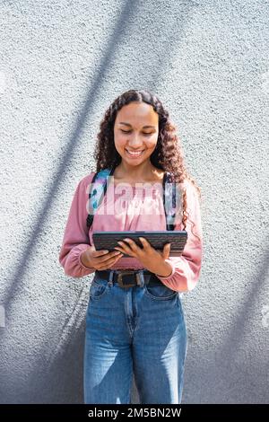 Latin university student woman smiling and using a tablet leaning against a wall Stock Photo