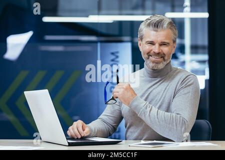 Portrait of senior man inside office, mature businessman with beard smiling and looking at camera, boss working at desk using laptop at work. Stock Photo