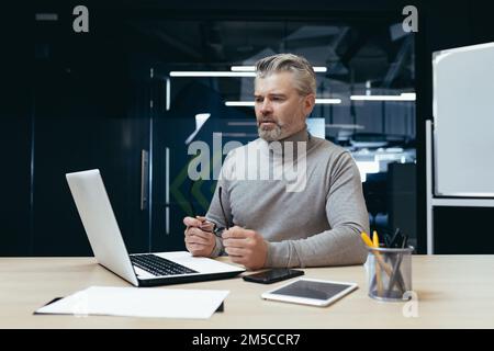 Serious thinking businessman inside office at work with laptop, senior gray haired man working sitting at desk. Stock Photo