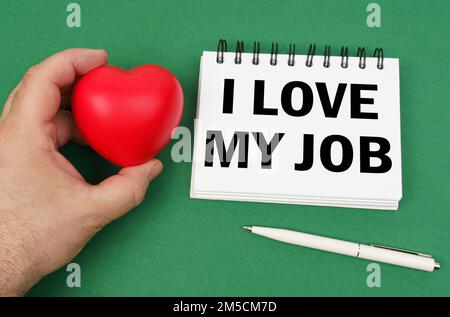 On the green surface are a pen, a notepad with the inscription - I love my job. A man has a heart in his hands. Business concept and symbols. Stock Photo