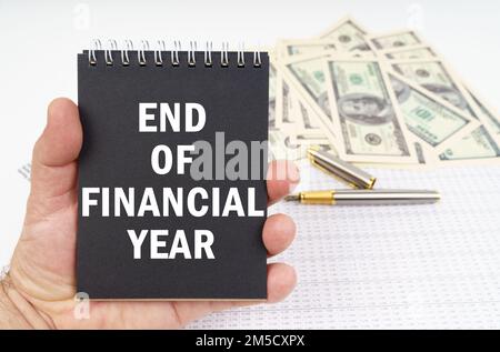 Business concept. In the man's hand is a notepad with the inscription - END OF FINANCIAL YEAR, in the background a pen and dollars. Pen and dollars bl Stock Photo