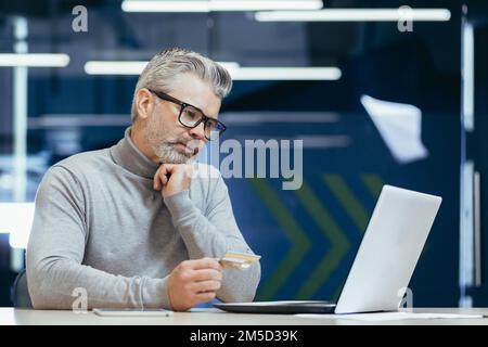 Senior gray haired businessman in office frustrated and sad trying to make bank transaction and purchase in online internet store, man holding bank credit card and using laptop. Stock Photo