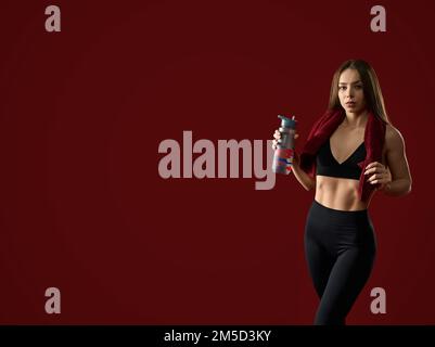 Front view of female fitness trainer after training with towel around neck holding shaker with water. Attractive brunette woman in black posing with protein drink in red studio, looking at camera. Stock Photo