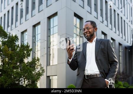 Successful and satisfied african american businessman using phone outside modern office building, mature man in business suit smiling and looking at smartphone screen texting. Stock Photo