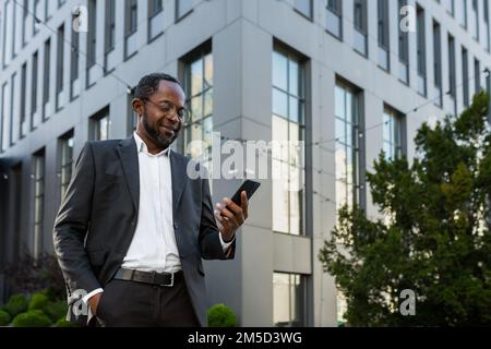 Successful and satisfied african american businessman using phone outside modern office building, mature man in business suit smiling and looking at smartphone screen texting. Stock Photo