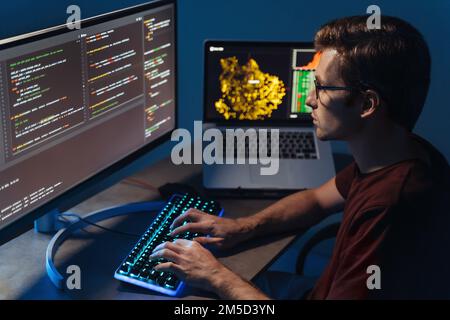 Profile closeup view of young handsome program developer writing software on multiple computer screens at home office. IT specialist, data scientist, coding concept Stock Photo