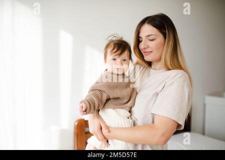 Cute infant baby girl under 1 year old on mother hands wear casual knit sweater and pants in bedroom. Look at camera. Motherhood. Stock Photo