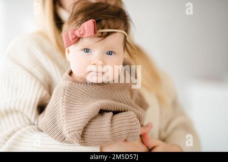 Cute infant baby girl under 1 year old on mother hands wear knit sweater and pants in bedroom. Look at camera. Motherhood. Stock Photo